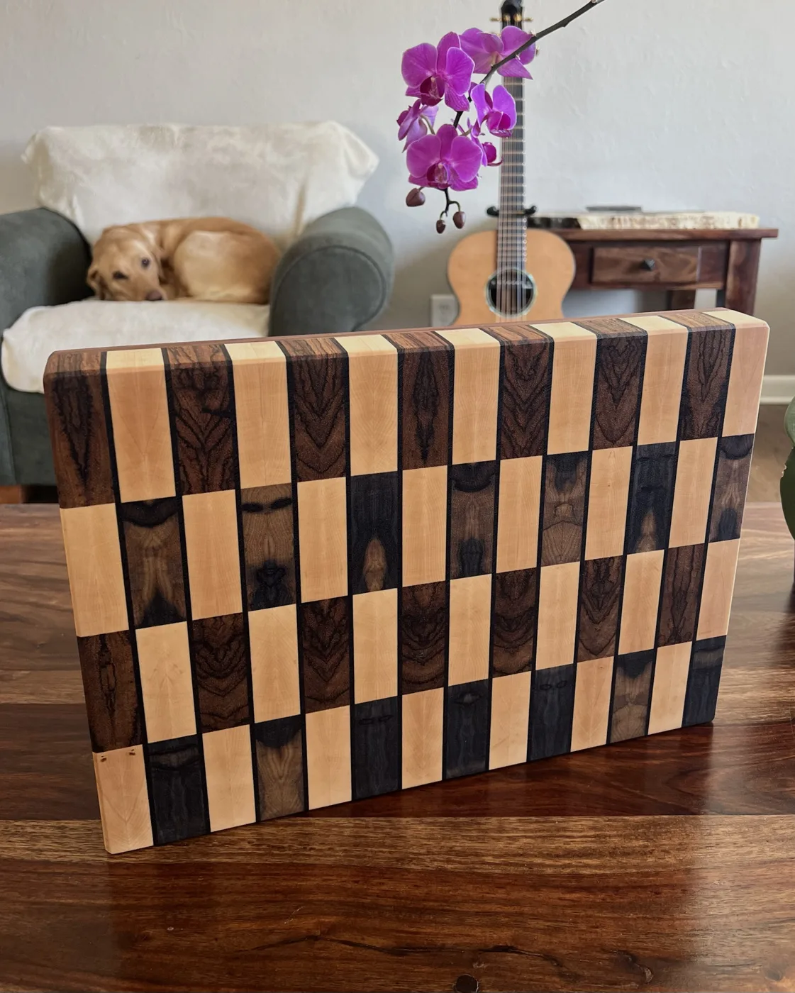 End-grain cutting board in alternating strips of maple and dark figured hardwood, with Zelda curled on a chair and a guitar in the background