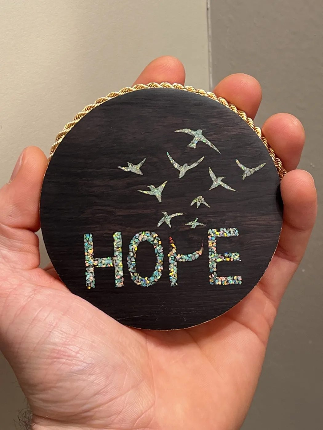 Round dark wood plaque held in a hand, inlaid with the word HOPE and a flock of birds in crushed shimmering stone, edged with gold rope