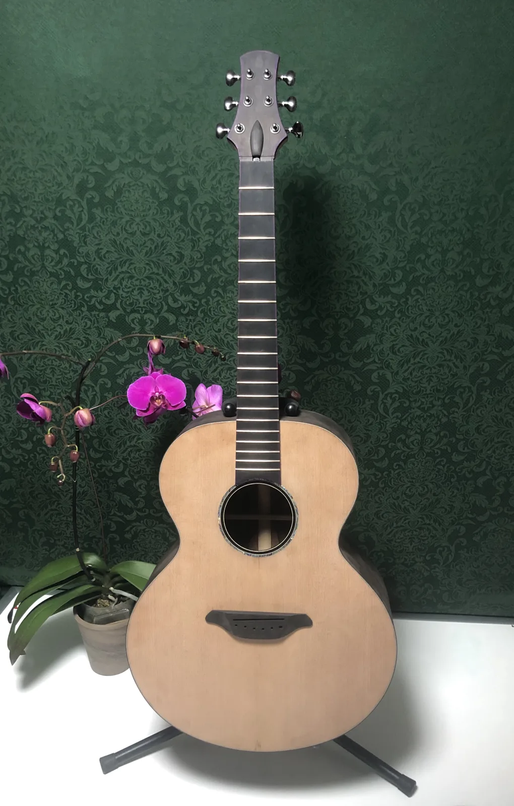 Build No. 001 before its finish, standing on a stand against a green damask wall beside a pink orchid, with the headstock tuners fitted