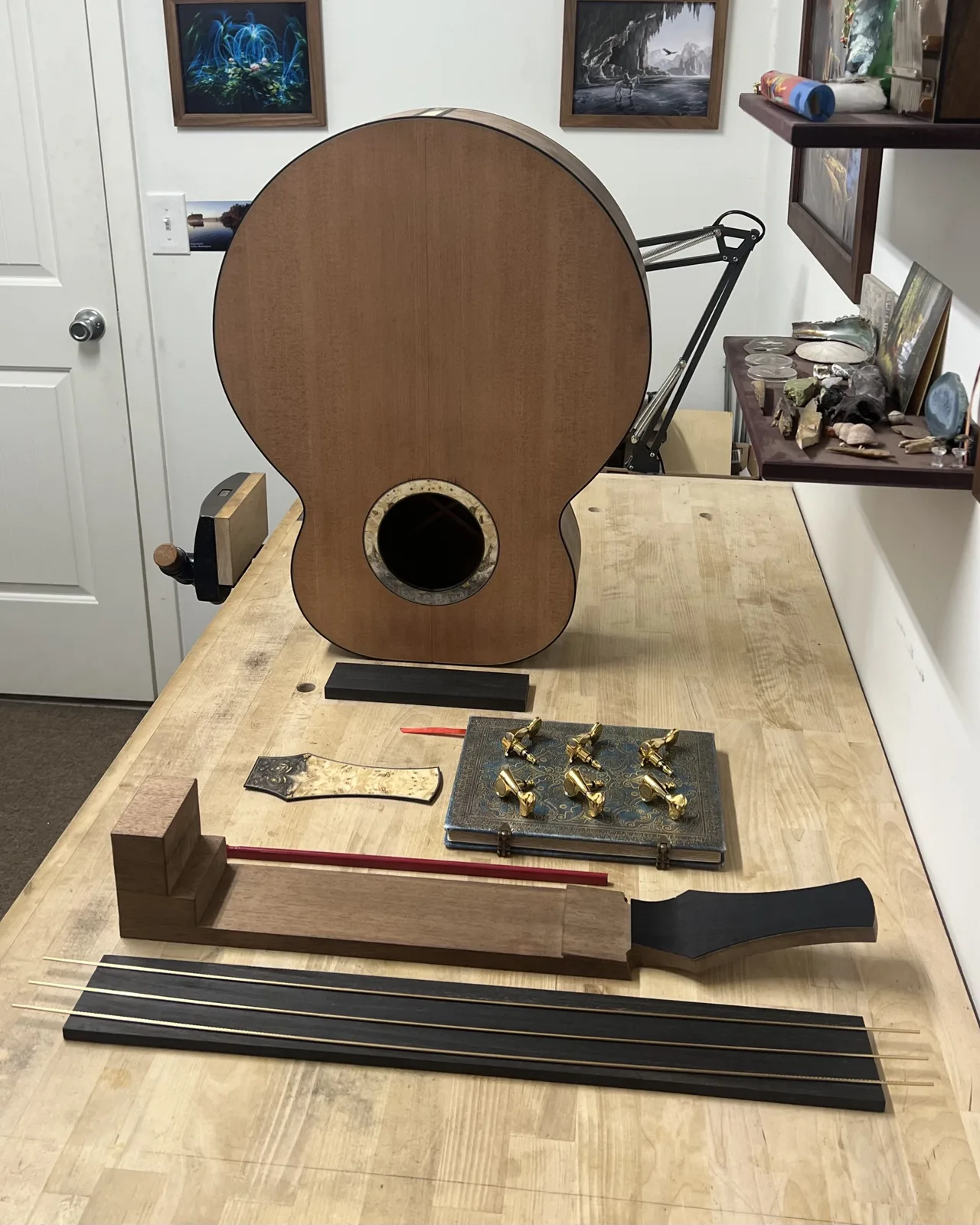 Build No. 002 in progress on the workbench: the closed body with a cedar top and burl rosette, the neck blank with its headstock, an ebony fretboard, fret wire, a burl headstock veneer and a set of gold tuners