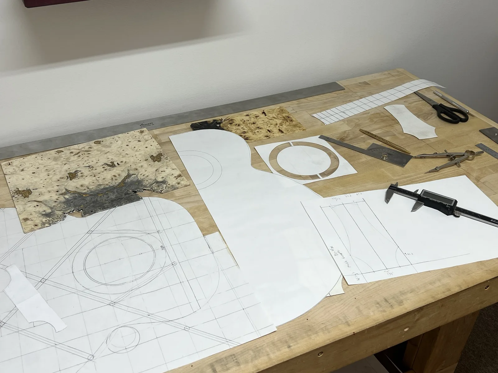 Full-size plans and templates on the bench: bracing layout, body outline and rosette, with burl veneer, a caliper and a compass