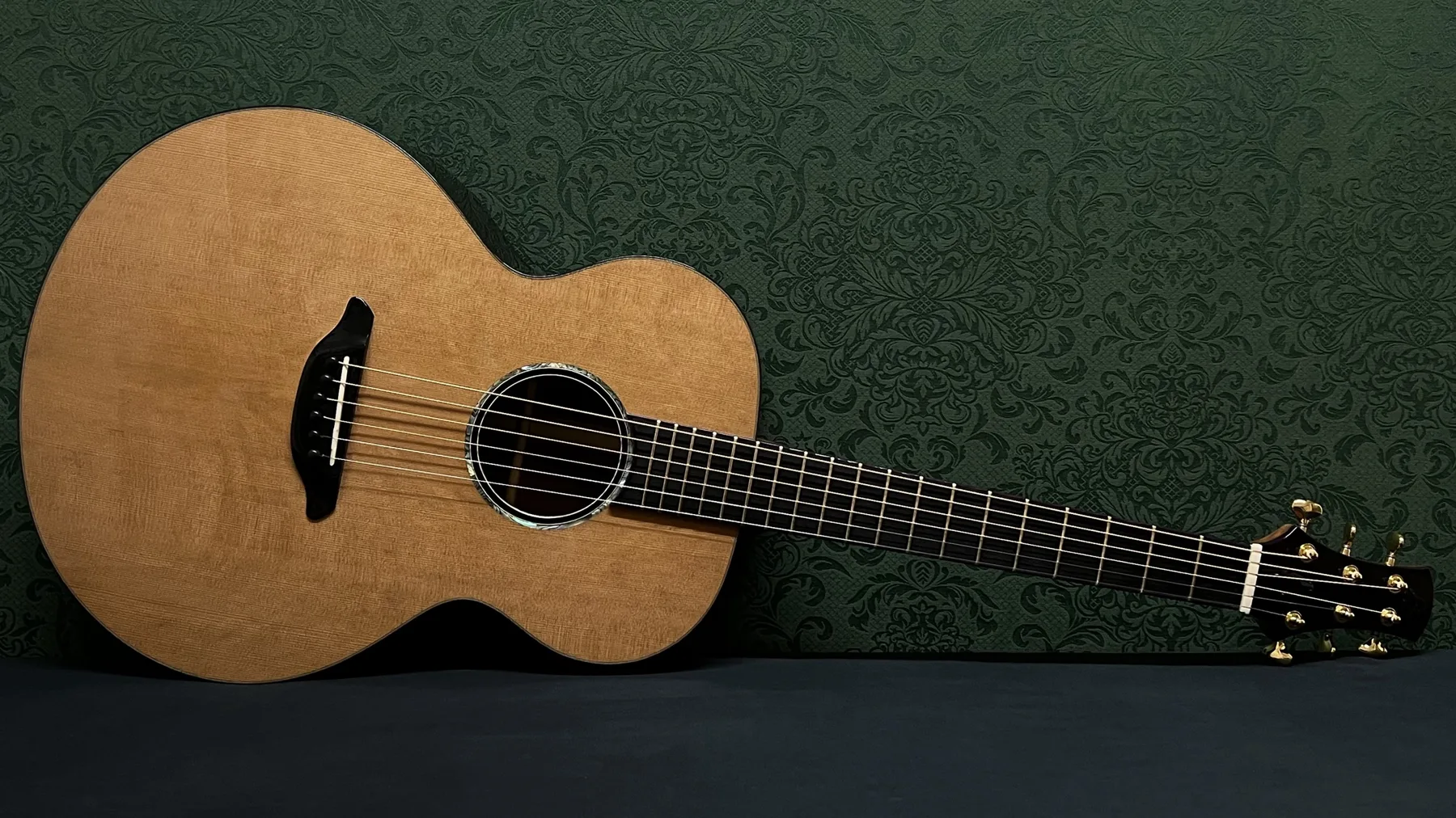 Build No. 001, a handbuilt acoustic guitar with a cedar top, leaning against a dark green damask wall