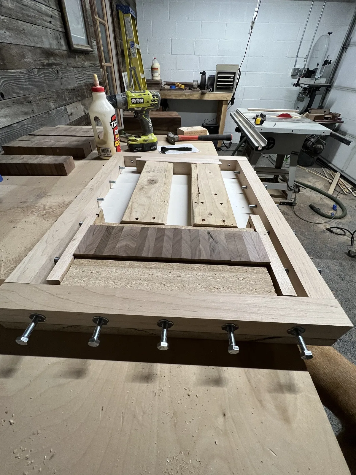 An end-grain walnut board glued up in a shop-made bolt clamping jig on the bench, with a table saw and band saw behind