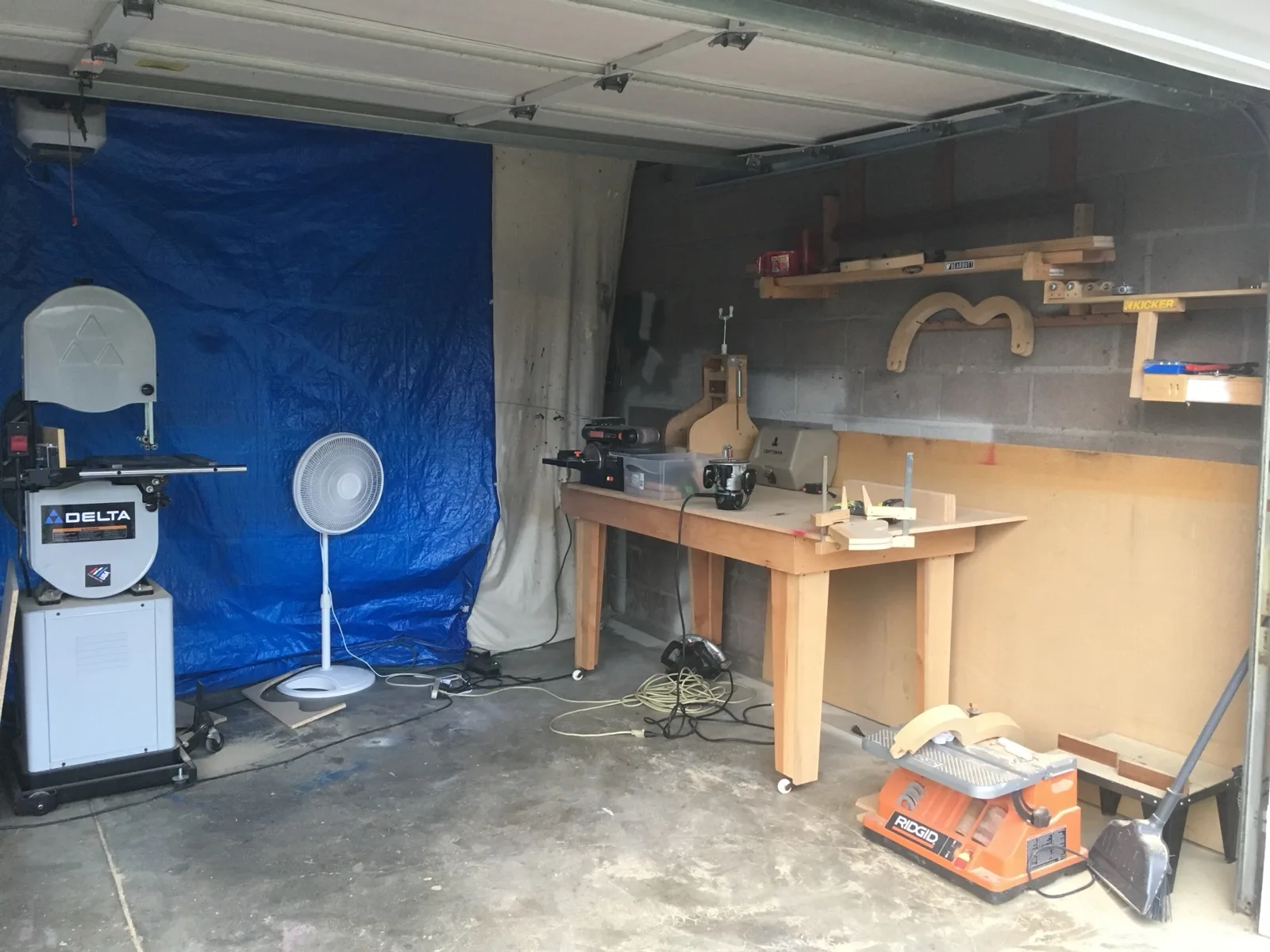 The Denver garage shop: a band saw, a wooden workbench with a sander and router, guitar molds and jigs on the shelves, and a blue tarp hung as a wall