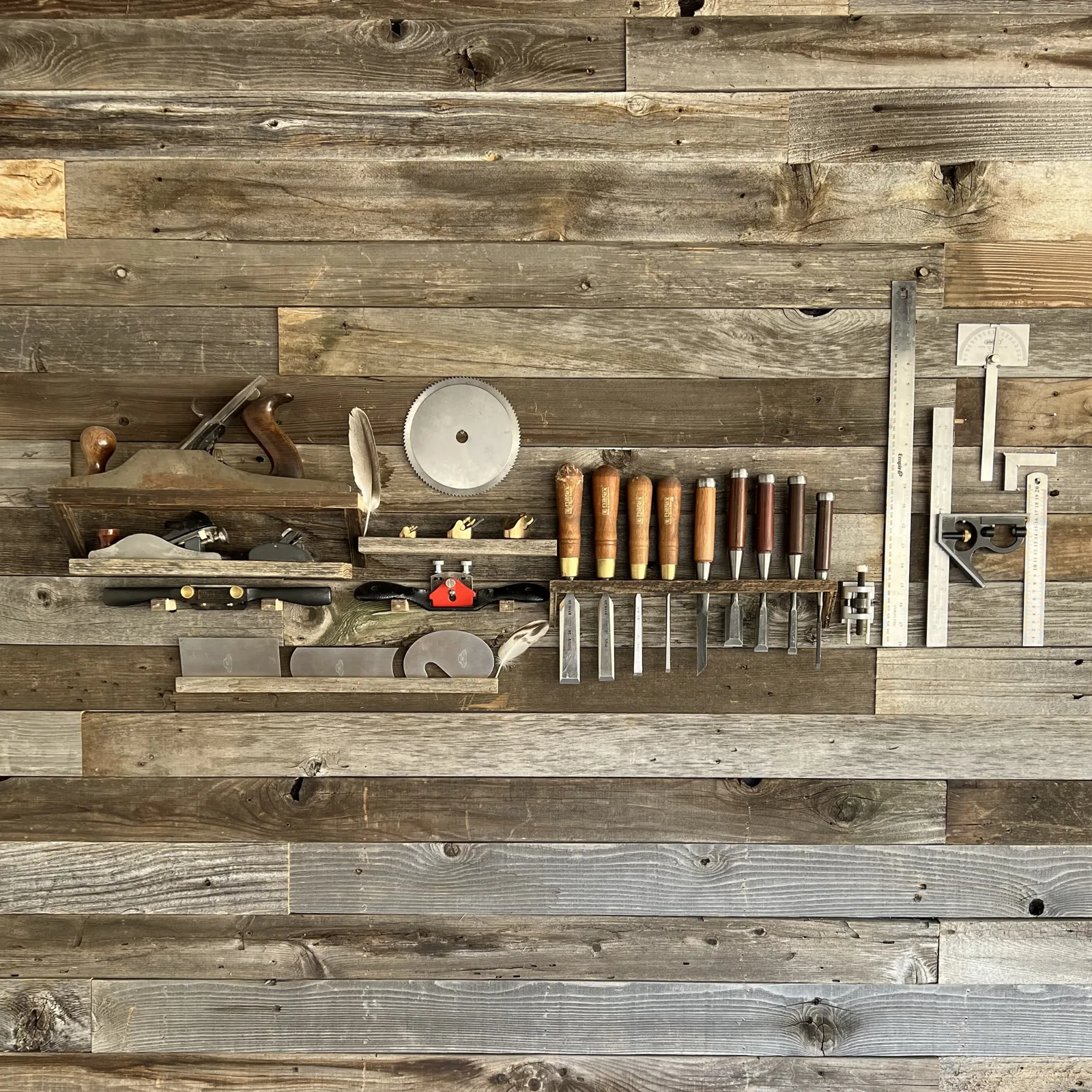 Hand tools arranged on a reclaimed barnwood wall: hand planes, spokeshaves, cabinet scrapers, a row of chisels, rulers and squares, with a couple of feathers tucked among them