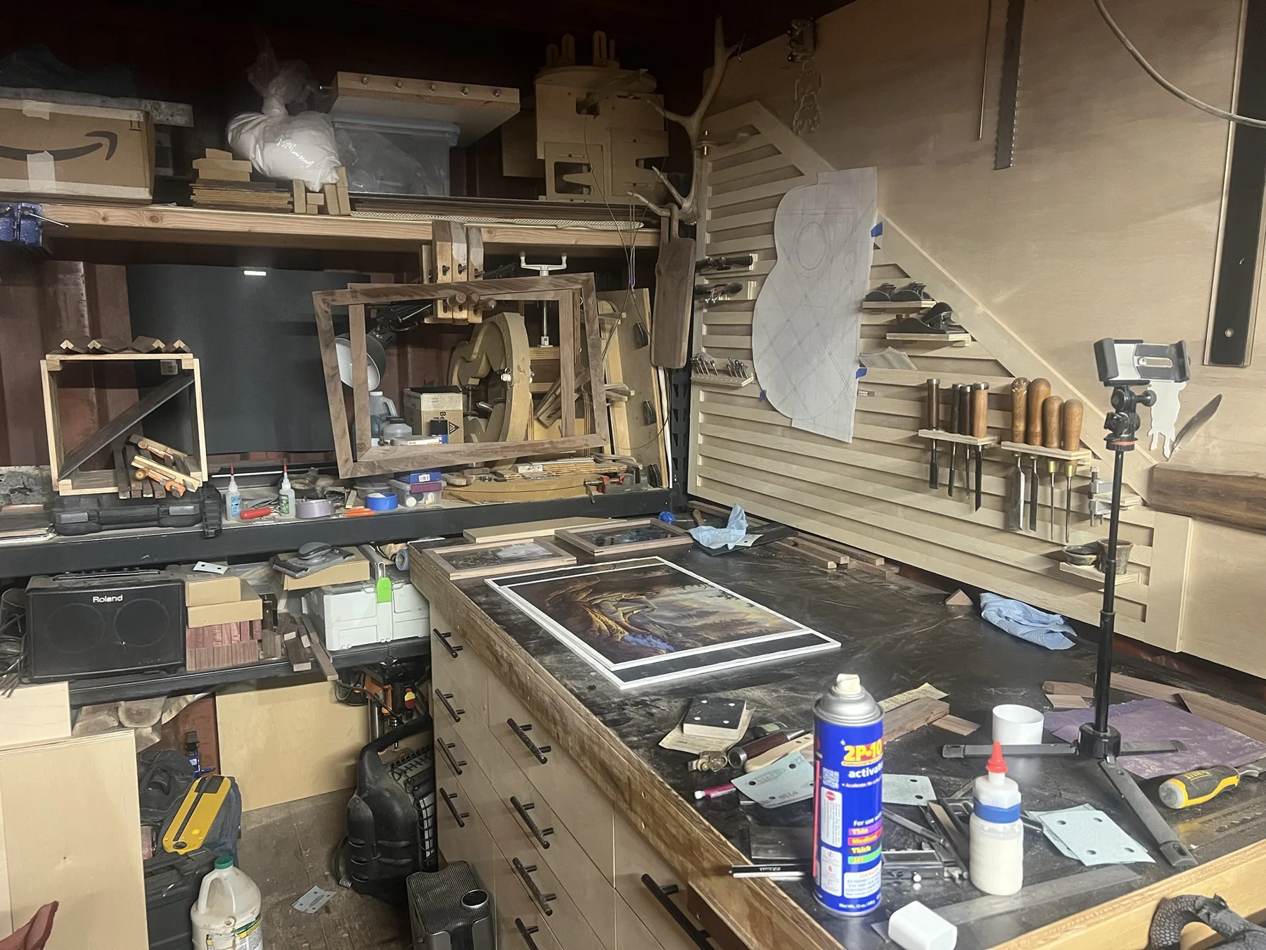 The Seattle shop: a long workbench with framing projects, guitar molds and jigs on the shelves, a slatted tool wall with chisels and planes, and a full-size guitar plan pinned up
