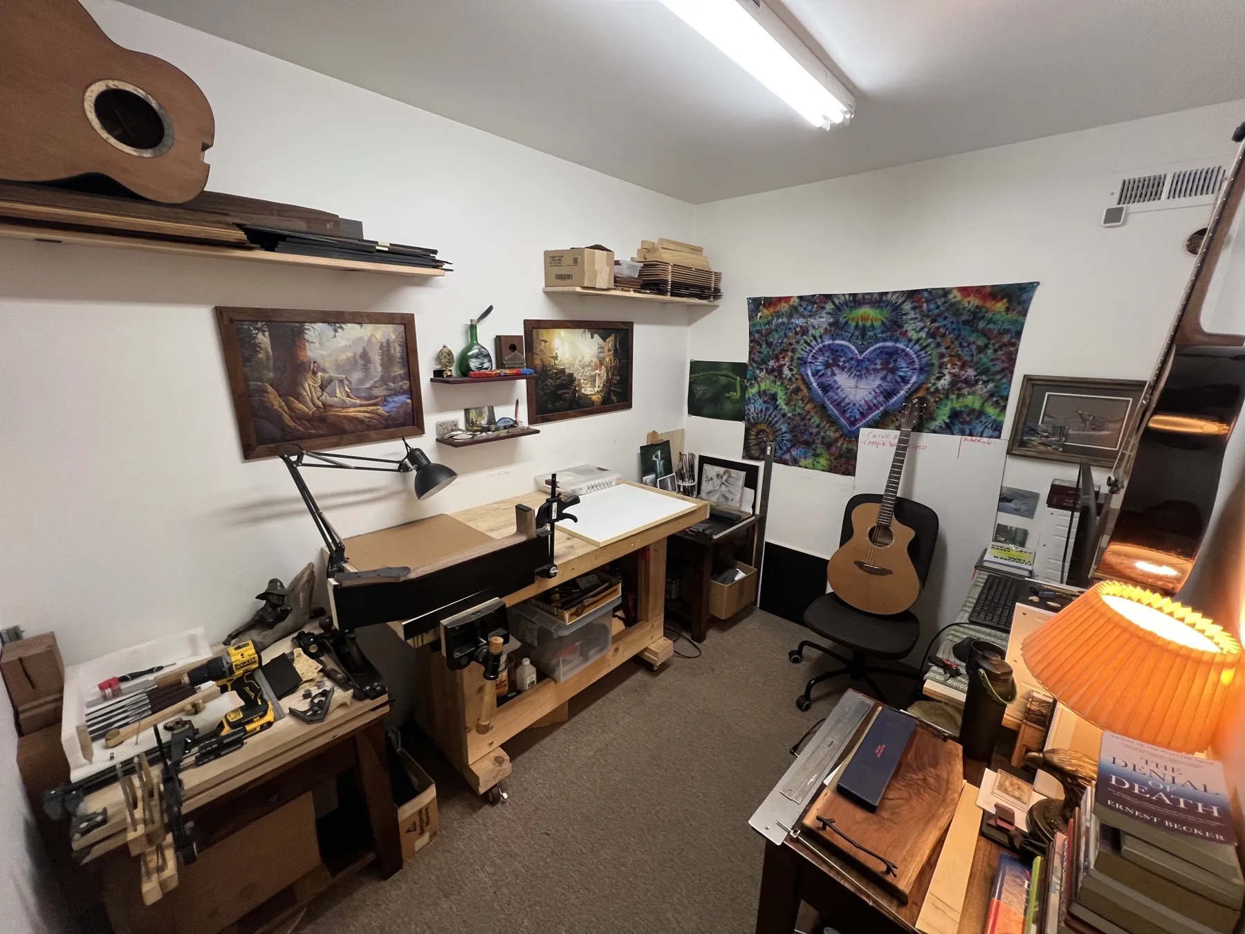 Brent's studio in Seattle: a wooden workbench with a neck in the vise, hand tools and clamps, wood stored on shelves above, framed paintings, a tie-dye heart tapestry and an acoustic guitar resting on a chair